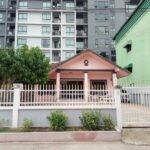 Front view of a small pink house with a peaked roof and white fence, set against a tall apartment block backdrop in Soi Khao Talo Pattaya