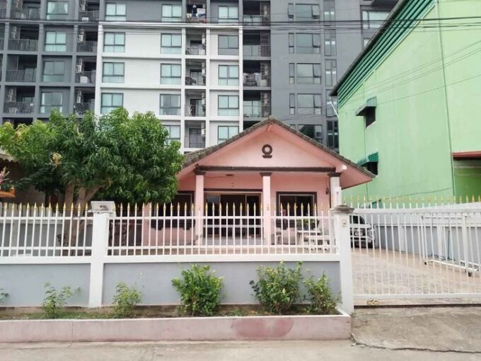 Front view of a small pink house with a peaked roof and white fence, set against a tall apartment block backdrop in Soi Khao Talo Pattaya