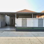 Modern single-story house with white walls, gray stone panels, and a covered carport; tiled driveway and small garden bed.