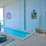 Indoor balcony pool on blue walls with mosaic edge, modern seating, and ocean view beyond the railing.