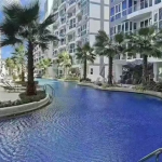Curved blue pool flanked by white apartment buildings and palm trees, with lounge chairs along the edge.