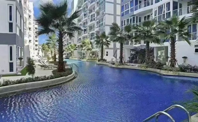 Curved blue pool flanked by white apartment buildings and palm trees, with lounge chairs along the edge.