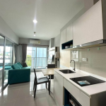 Open-plan apartment: white kitchen with sink and stove, a small dining table, teal sofa in the living area, and a balcony view.